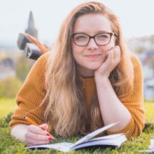 young woman laying on her belly on a field, journaling, with blurry cityscape in the background